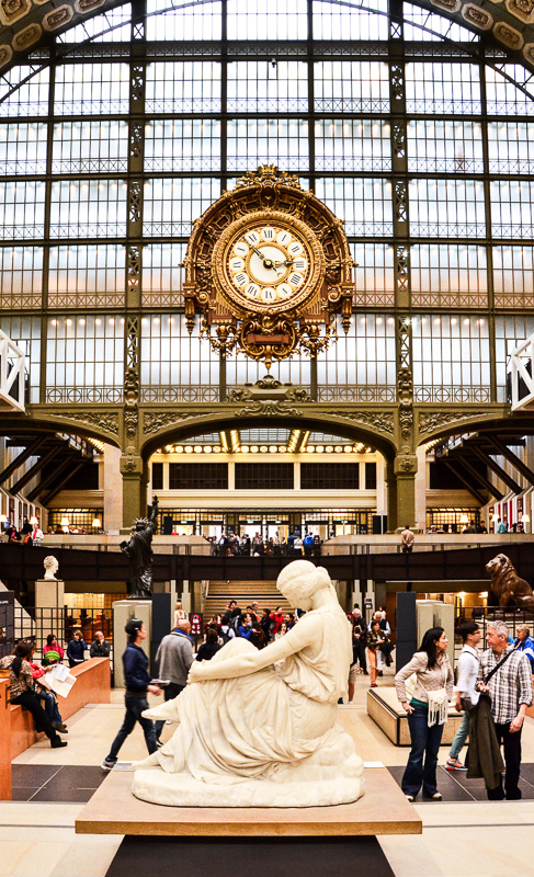 Visitors looking at white sculpture of woman with clock in the background on glass domed ceiling and walls in the Musee d'Orsay